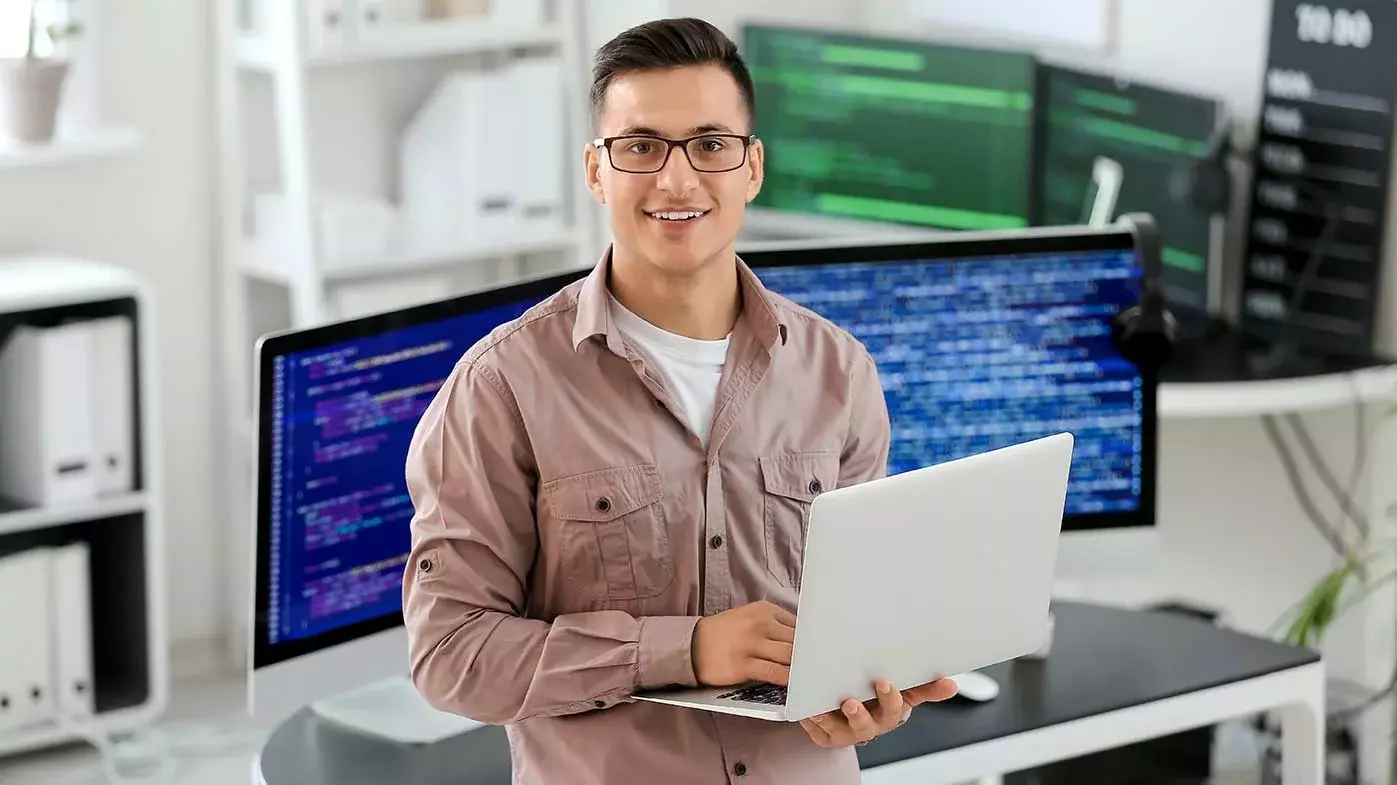Ein junger Mann steht in einem modernen Büro vor einem weißen Schreibtisch mit dunkler Schreibtischplatte. Er hält einen grauen Laptop in der Hand und blickt in die Kamera: Im Hintergrund sind zwei große Monitore zu sehen, auf denen Code verschwommen sichtbar ist. Im Hintergrund sind weitere weiße Büromöbel zu sehen und auch zwei weitere große Monitore, auf denen Code verschwommen sichtbar ist.  
