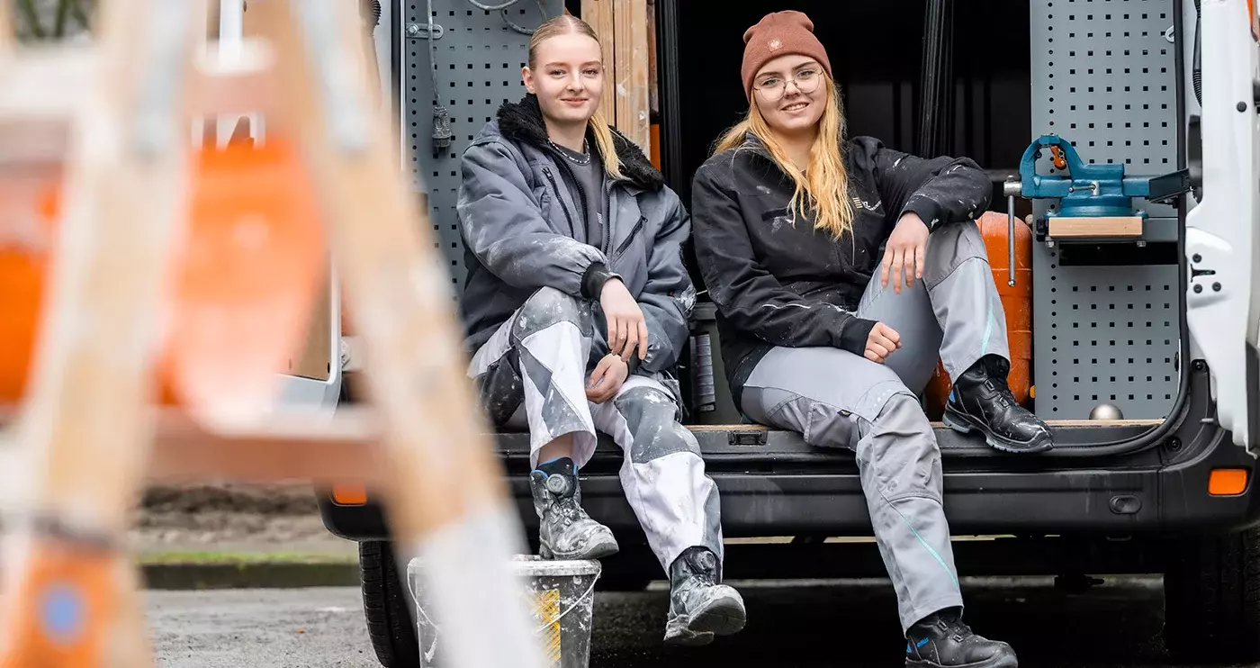 Zwei junge Frauen sitzen auf der Ladefläche eines kleinen Transporters. Sie tragen beide Handwerkerkleidung. Im Hintergrund ist diverses Werkzeug im Auto zu sehen. Sie schauen und lächeln beide in die Kamera. Im Vordergrund ist unscharf eine Leiter zu sehen