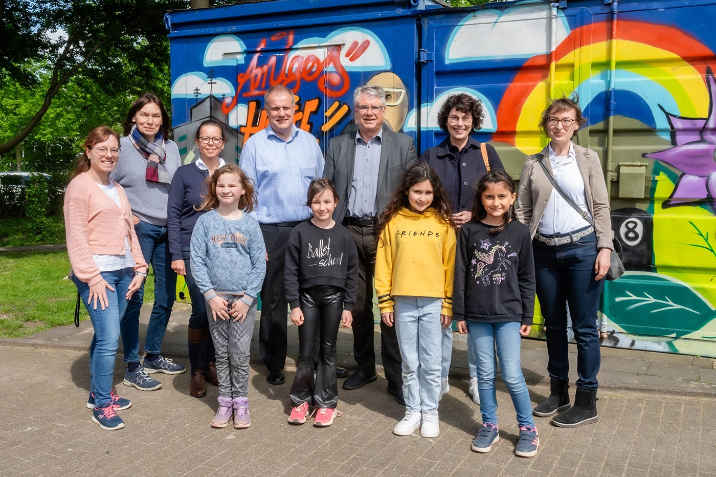Auf dem Bild sind sieben Erwachsene zu sehen, davon fünf Frauen und zwei Männer. Vor ihnen stehen vier Mädchen. Sie schauen und lächeln alle in die Kamera. Im Hintergrund ist ein mit Graffiti besprühter Container zu sehen