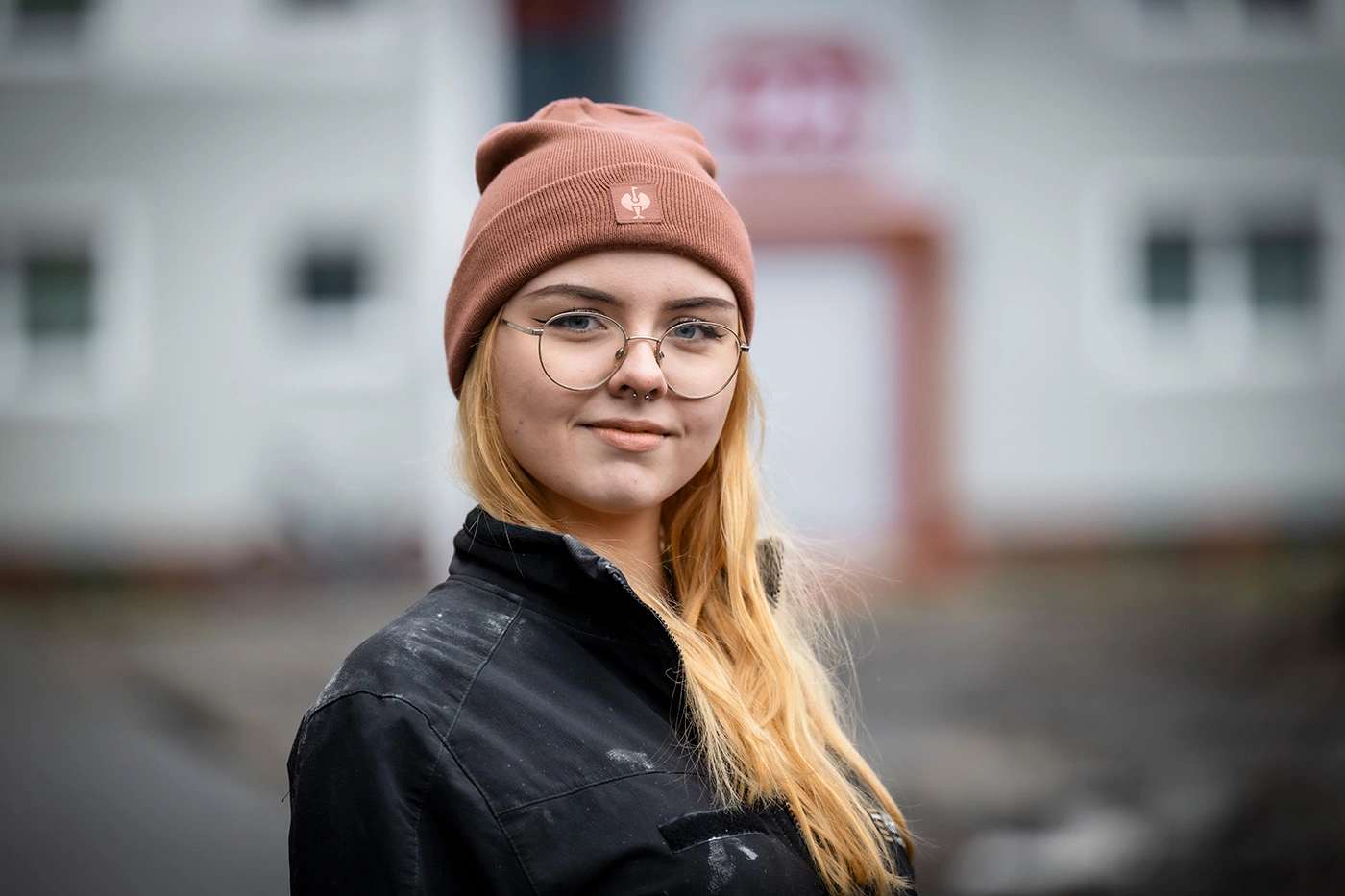 Junge Frau steht leicht seitlich und lächelt in die Kamera. Sie trägt eine Brille, hat ein Septum und sie trägt eine ockerfarbene Mütze und eine schwarze Jacke. Ihre rötlich blonden Haare trägt sie offen