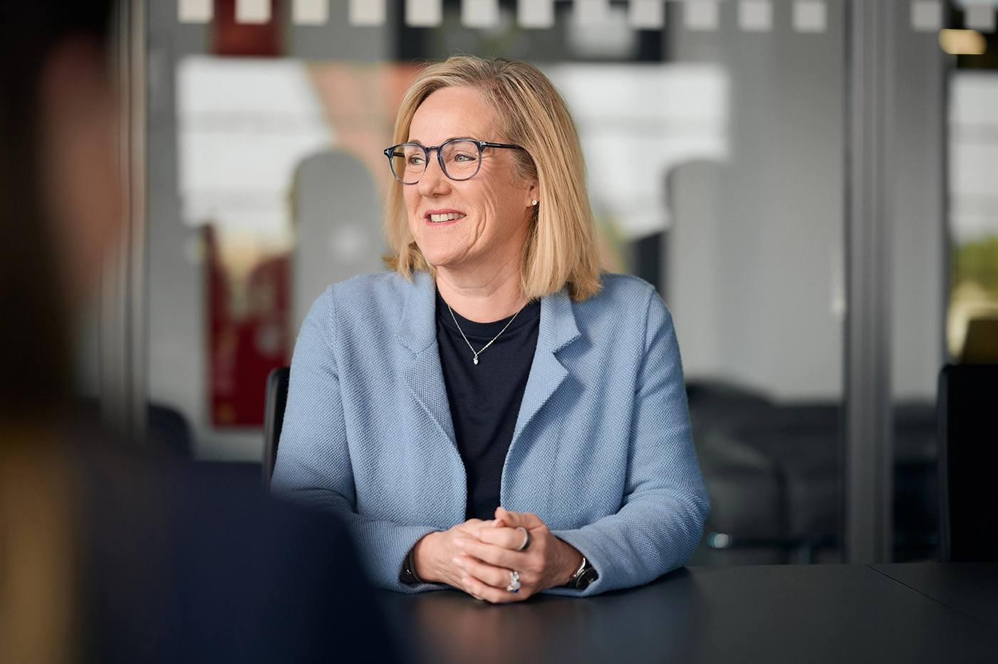 Eine blonde Frau mit Brille sitzt an einem schwarzen Tisch. Man sieht sie im Profil. Sie trägt eine schwarze Bluse und einen hellblauen Blazer