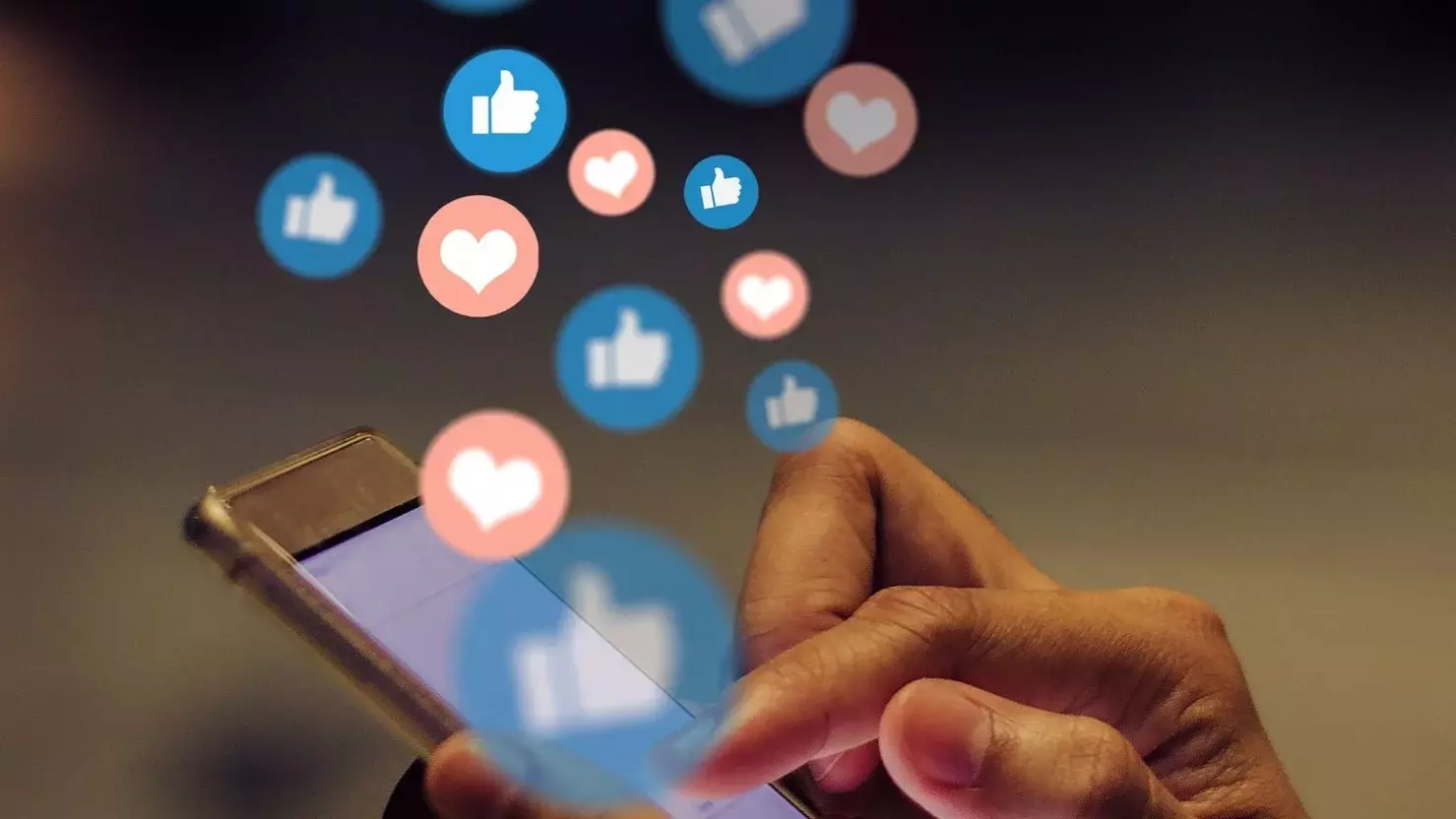 Ein Mensch tippt auf den Bildschirm eines Smartphones. Über dem Smartphone steigen viele blaue Buttons auf, die das weiße Facebook Gefällt mir-Symbol abbilden. Es steigen außerdem Buttons über dem gerät auf, die sie den Instagram Like-Symbolen ähneln. Es handelt sich um ein weißes Herz auf rotem Hintergrund.
