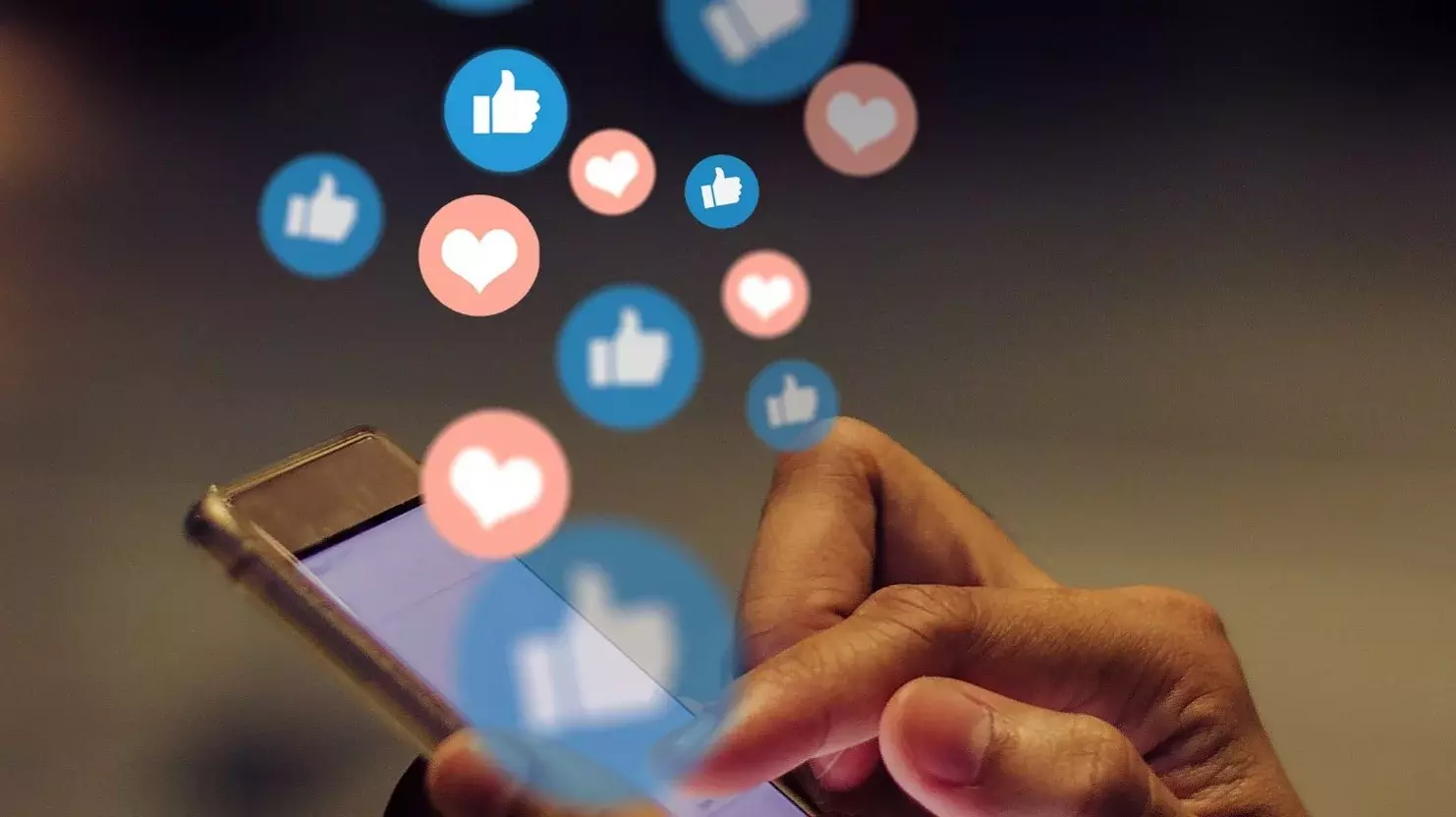 Ein Mensch tippt auf den Bildschirm eines Smartphones. Über dem Smartphone steigen viele blaue Buttons auf, die das weiße Facebook Gefällt mir-Symbol abbilden. Es steigen außerdem Buttons über dem gerät auf, die sie den Instagram Like-Symbolen ähneln. Es handelt sich um ein weißes Herz auf rotem Hintergrund.