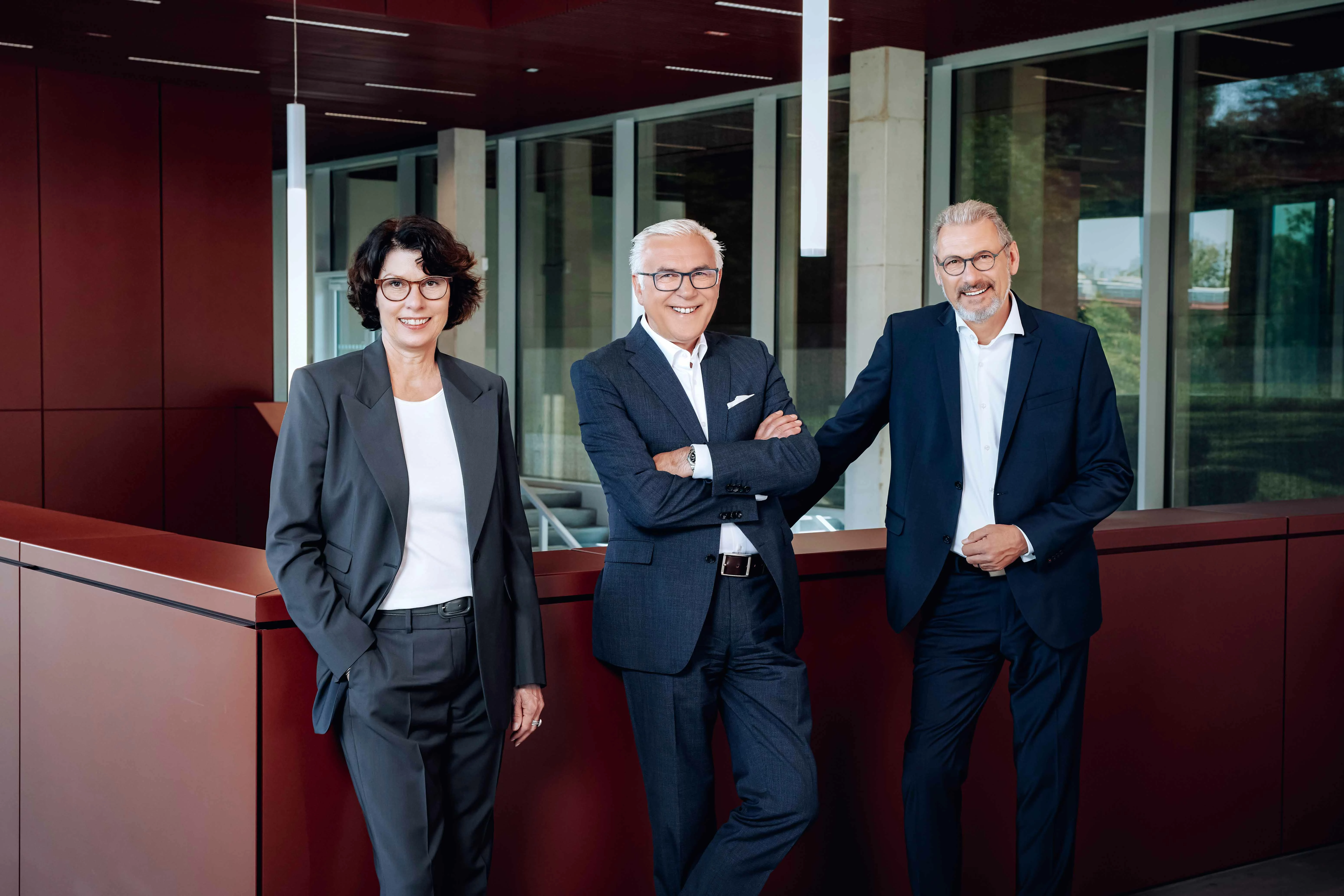Die drei Mitglieder der VIVAWEST-Geschäftsführung sind gemeinsam im Foyer des Kesselhauses am VIVAWEST-Campus abgebildet. Von links: Arbeitsdirektorin Karin Erhard, Haluk Serhat und Uwe Eichner, der Vorsitzende der Geschäftsführung. Alle drei tragen dunkle Anzüge und weiße Hemden, beziehungsweise Blusen. Sie lehnen an einer rostroten Geländer.
