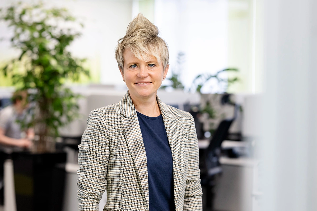 Eine Frau ist im Porträt zu sehen. Sie hat kurze blonde Haare, die teilweise nach oben toupiert sind. Sie trägt ein blaues T-Shirt und einen braun-schwarz-beige-karierten Blazer. Sie lächelt leicht und schaut in die Kamera. Im Hintergrund sind Tische und eine Pflanze zu sehen.