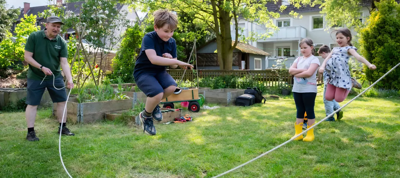 Auf dem Bild sind vier Kinder und ein Mann in einem Garten zu sehen. Der Mann schwingt ein Seil und ein Junge springt darüber. Ein Mädchen steht da und beobachtet die Szene das Mädchen daneben springt ebenfalls in die Luft. Im Hintergrund sind Beete und ein Bollerwagen zu sehen. Der zweite Junge läuft im Hintergrund