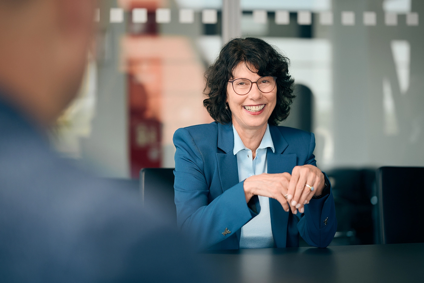 Eine Frau mit dunklen, kinnlangen Haaren und einer Brille sitzt an einem schwarzen Schreibtisch und lächelt in die Kamera. Sie trägt eine hellblaue Bluse und einen dunkelblauen Blazer