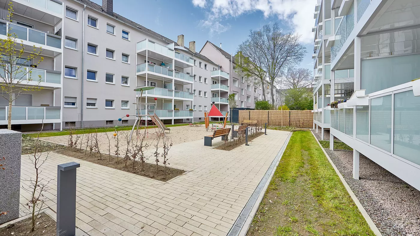 Modernes Wohnquartier mit Mehrfamilienhäusern in grau. An den Häusern sind Balkone angebracht. In der Mitte ist ein Spielplatz mit Rutsche und Klettergerüst zu sehen. Es gibt einen Sandkasten, ein paar Sitzbänke und eine kleine Grünfläche