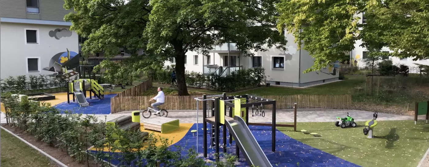 Ausschnitt eines VIVAWEST-Quartiers. Darauf ist ein Spielplatz zu sehen mit Rutschen, Kletterturm und Wippe. Umgeben ist der Spielplatz von Bäumen und niedrigen Hecken. Auf einem Weg fährt ein Mann auf einem Fahrrad am Spielplatz vorbei. Im Hintergrund ist zwischen den Bäumen ein weißes Haus zu erkennen.