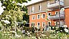 Garten, umgeben von Blumen und Bäumen. Im Garten steht eine Schaukel auf der zwei Kinder mit dem Rücken zur Kamera sitzen. Im Hintergrund ist ein orangenes Haus zu sehen