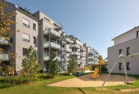 Innenhof-Ansicht des neu angekauften VIVAWEST-Quartiers Weserhöfe in Düsseldorf. Im Vordergrund siegt man eine Sandfläche, auf der eine gelbe Rutsche steht, rechts daneben steht eine Metallschaukel. Im Hintergrund sieht man die vierstöckigen Gebäude mit weißen Fassaden und Vorstellbalkonen. Copyright des Bildes: heck + schmiegel architektur 