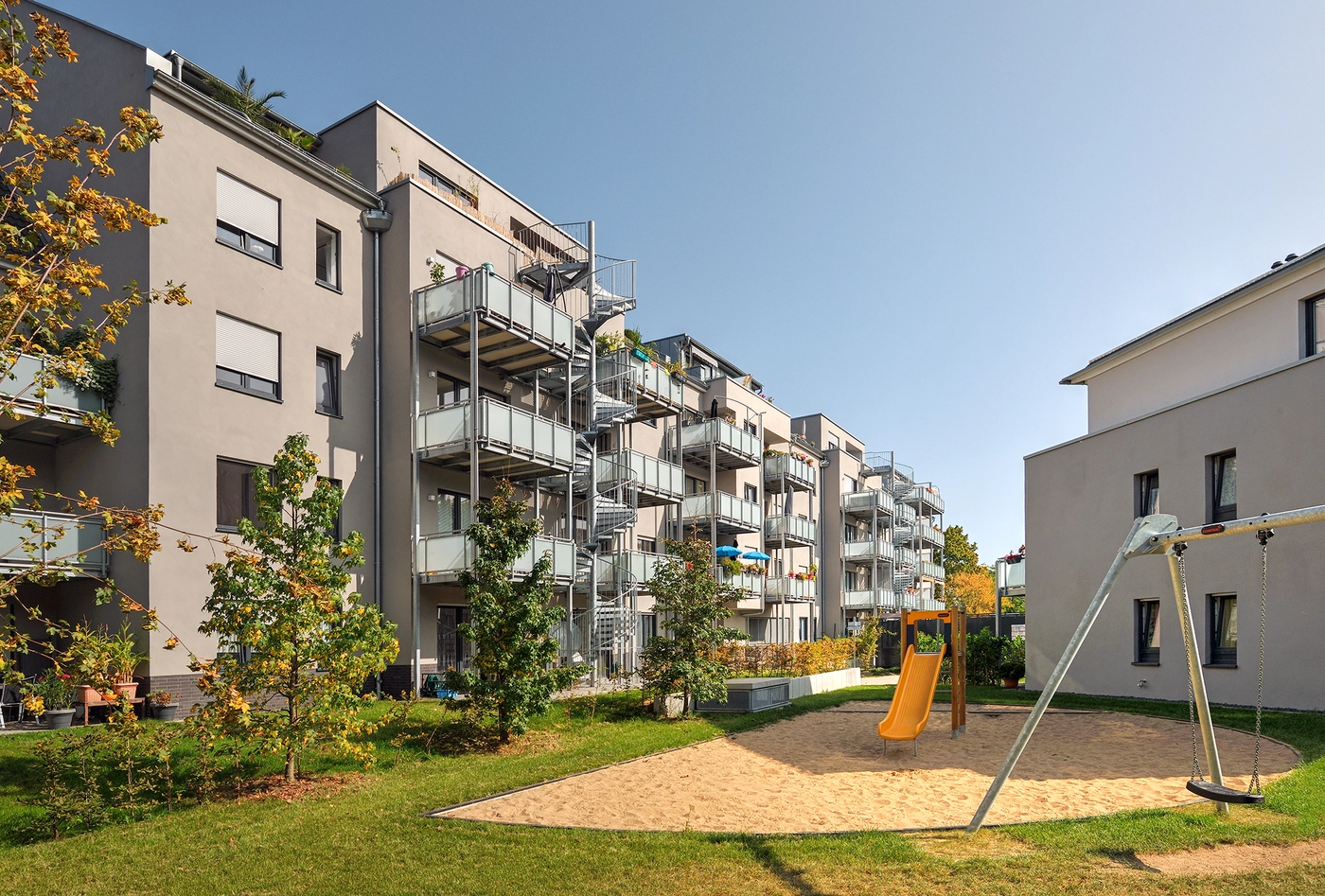 Innenhof-Ansicht des neu angekauften VIVAWEST-Quartiers Weserhöfe in Düsseldorf. Im Vordergrund siegt man eine Sandfläche, auf der eine gelbe Rutsche steht, rechts daneben steht eine Metallschaukel. Im Hintergrund sieht man die vierstöckigen Gebäude mit weißen Fassaden und Vorstellbalkonen. Copyright des Bildes: heck + schmiegel architektur 