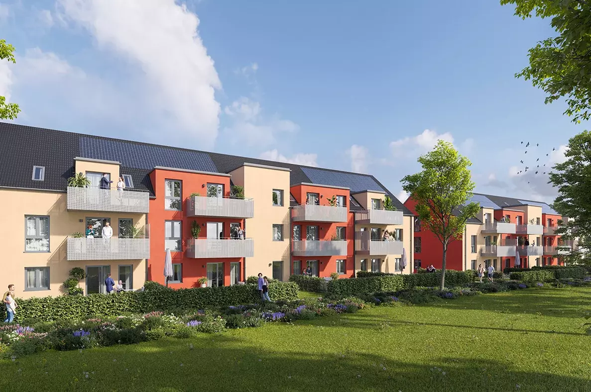 Zwei Mehrfamilienhäuser-Reihen stehen nebeneinander. Die Fassaden erstrahlen in einem dunklen Orange und in einem Eierschalengelb. An den Vorderseiten des Hauses sieht man mehrere Balkone mit Menschen darauf. Vor den Häusern befindet sich eine große Grünfläche mit kleinen Büschen und Blumen. Ein paar Menschen gehen vor den Häusern spazieren