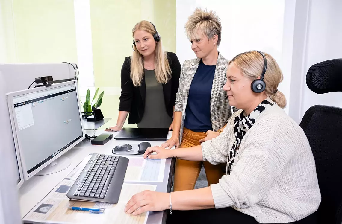 Drei Frauen befinden sich an einem Schreibtisch. Eine der Frauen sitzt auf einem Bürostuhl und zeigt den anderen beiden gerade etwas am PC. Alle drei schauen konzentriert auf den Bildschirm.