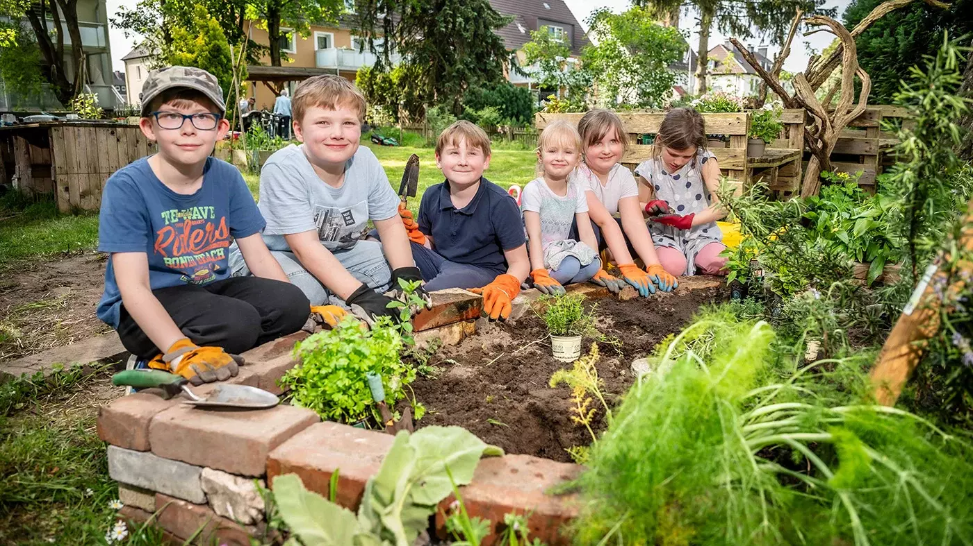 Auf dem Bild sind drei Jungs und drei Mädchen zu sehen, die an einem Kräuterbeet sitzen. Ein Kind hält eine Gartenschaufel in der Hand, sie tragen Gartenhandschuhe. Im Hintergrund ist Garten zu sehen
