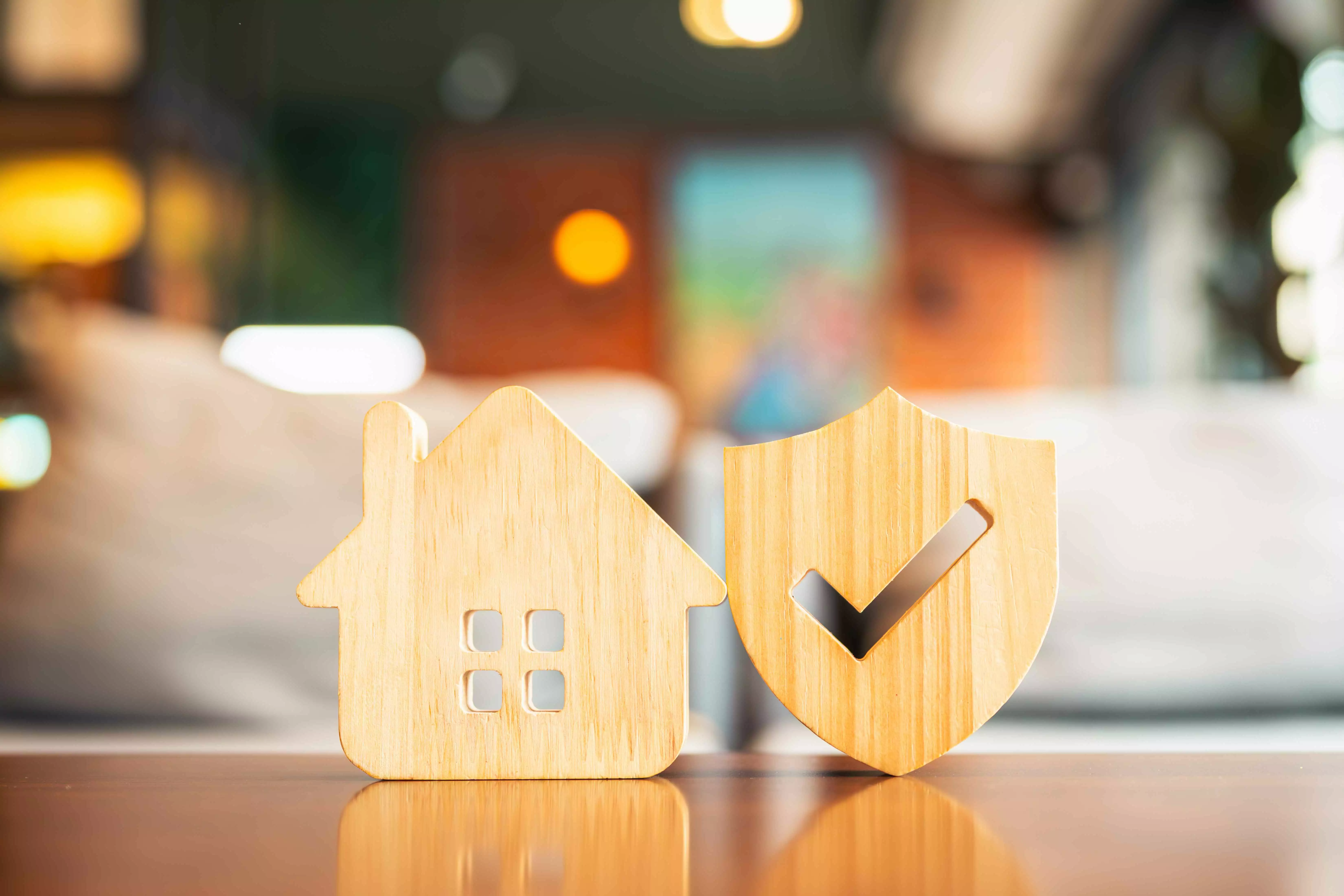 Zwei Symbole aus hellem Holz sind vor einem verschwommenen Hintergrund zu sehen. Das linke Symbol zeigt ein Haus und das rechte Symbol zeigt einen Schild, in dem ein ausgefrästes Häkchen. 
