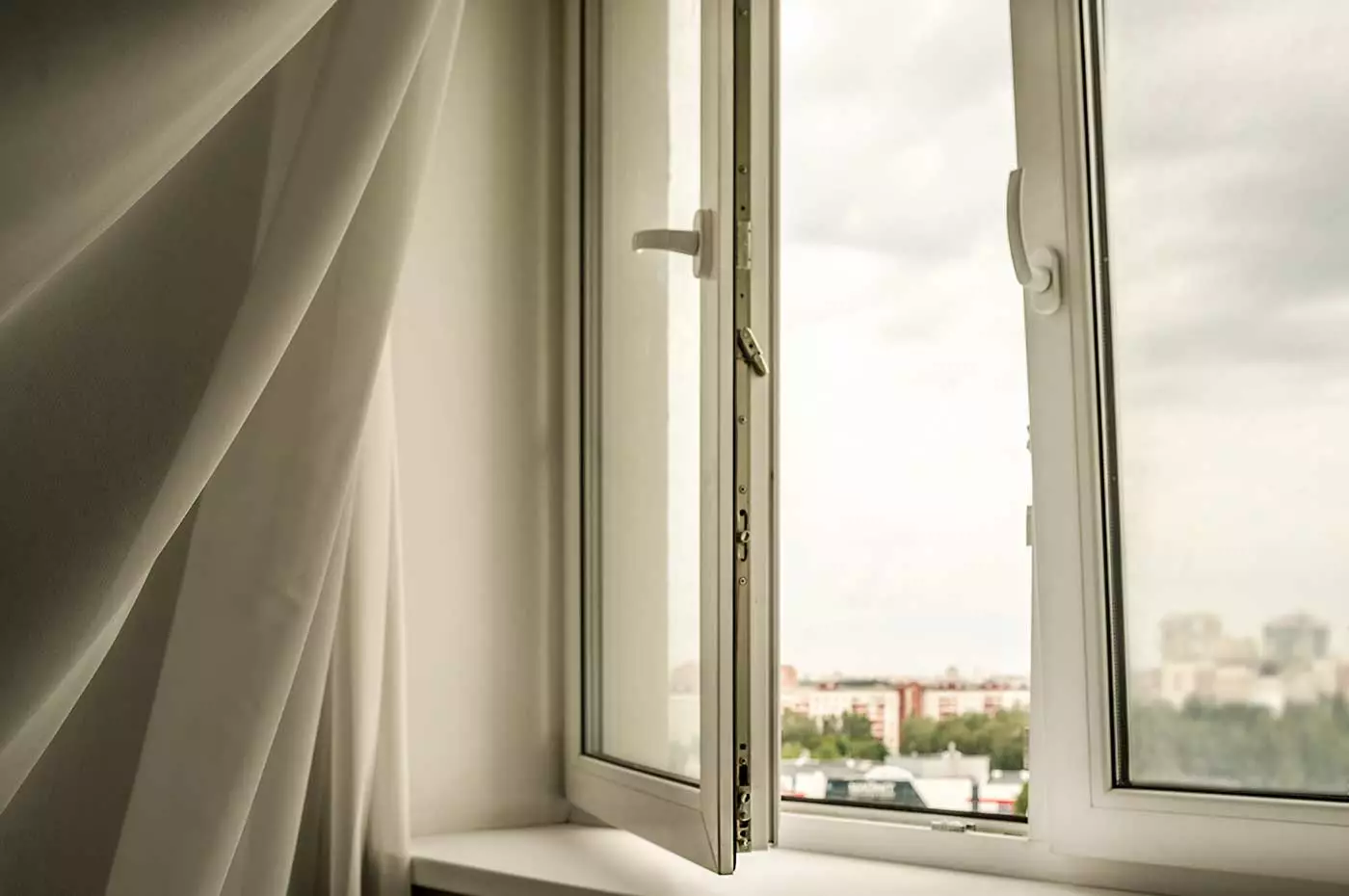 Ein geöffnetes Zimmer in einem Wohnraum. Man sieht einen weißen, transparenten Vorgang im Wind flattern. Im Hintergrund des Bildes ist verschwommen ein Stadtpanorama zu sehen.