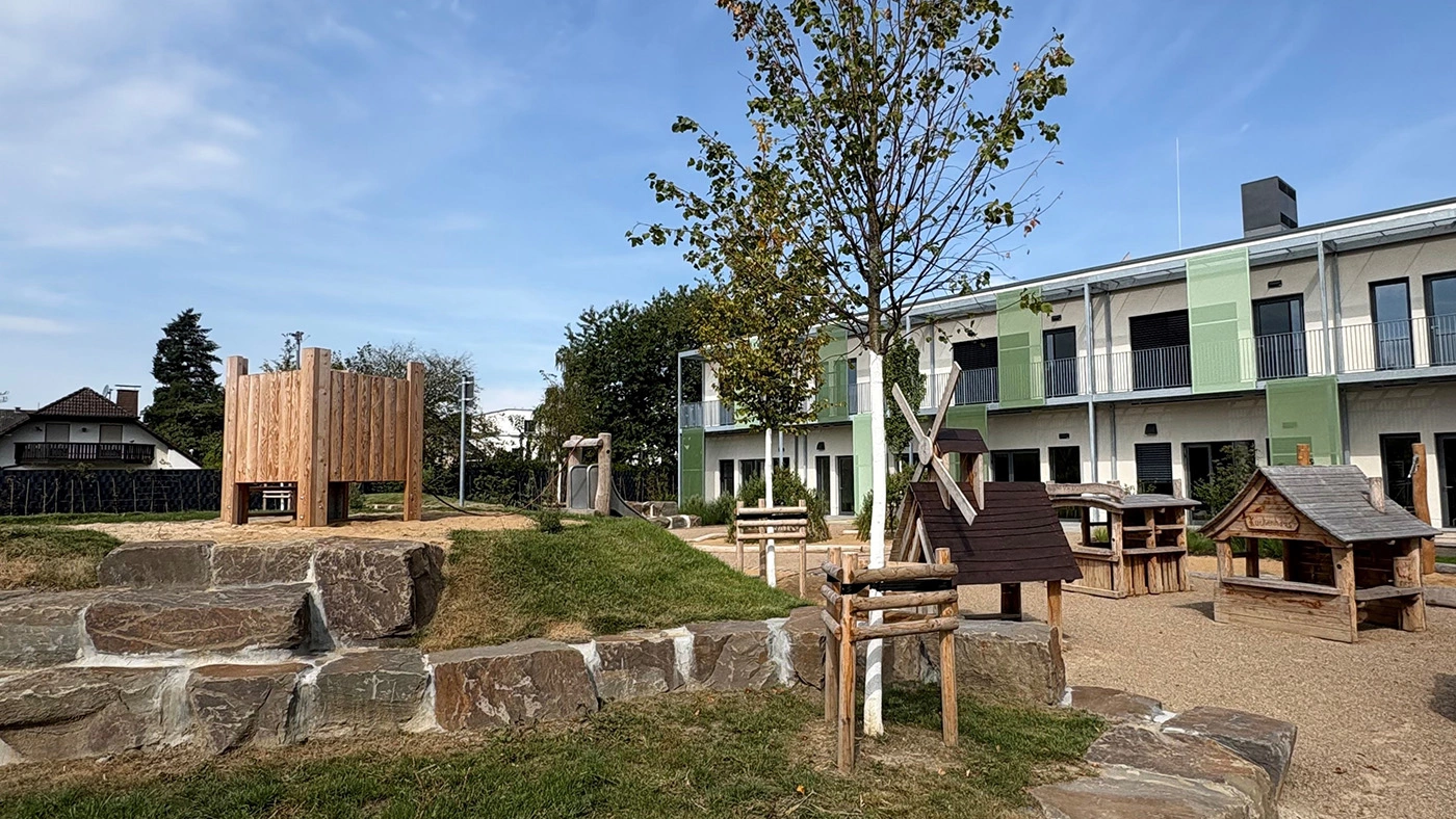 Spielplatz mit kleinen Holzhäuschen und einer Rutsche. Im Hintergrund steht ein grün-weißes Neubaugebäude