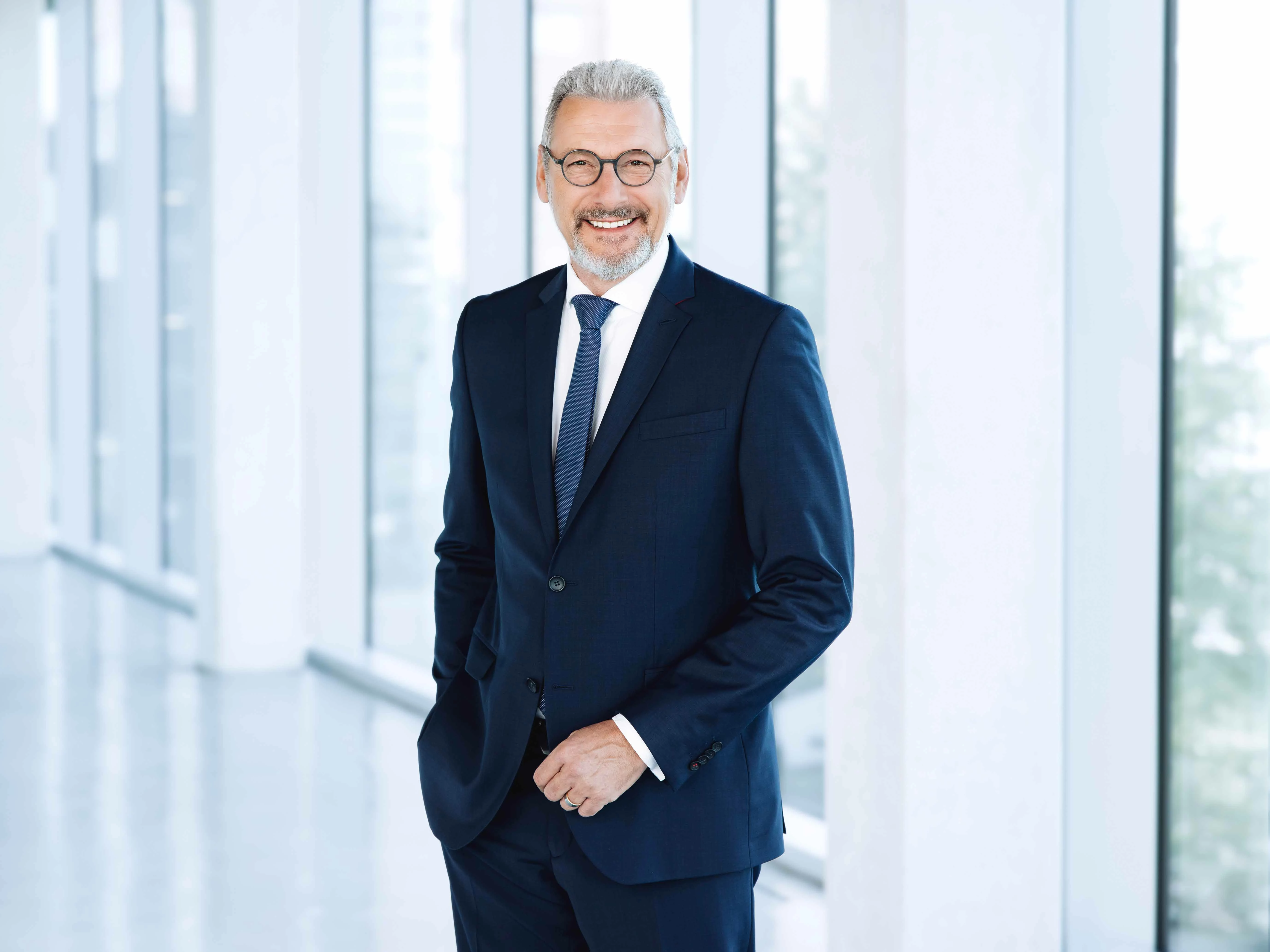 Uwe Eichner, der Vorsitzende der VIVAWEST-Geschäftsführung im Portrait. Er steht vor einer Fensterfront im Foyer des Kesselhauses am VIVAWEST-Campus und schaut lächelnd in die Kamera. Er trägt einen dunkelblauen Anzug, ein weißes Hemd und eine dunkelblaue Krawatte.
