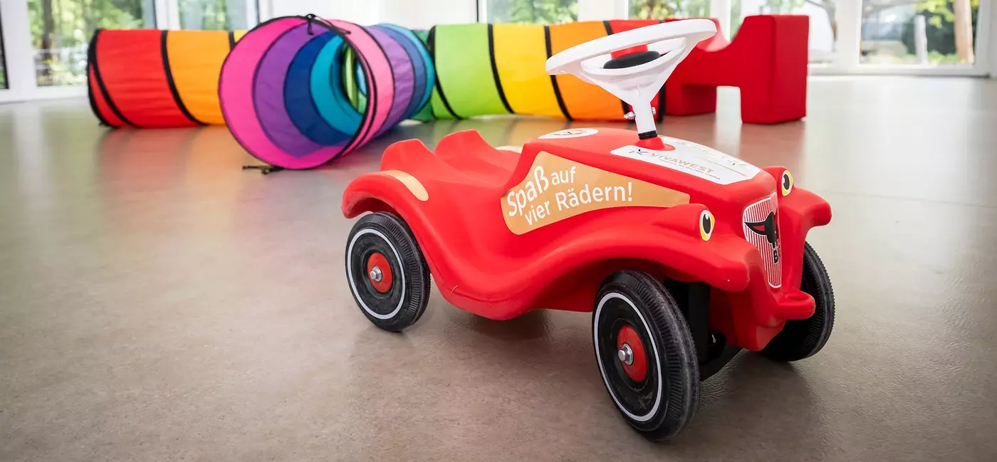 Ein rotes Bobbycar steht in einem Raum. Auf der Seite ist ein Sticker angebracht, auf dem "Spaß auf vier Rädern" steht. Im Hintergrund ist ein bunter Spieltunnel zu sehen