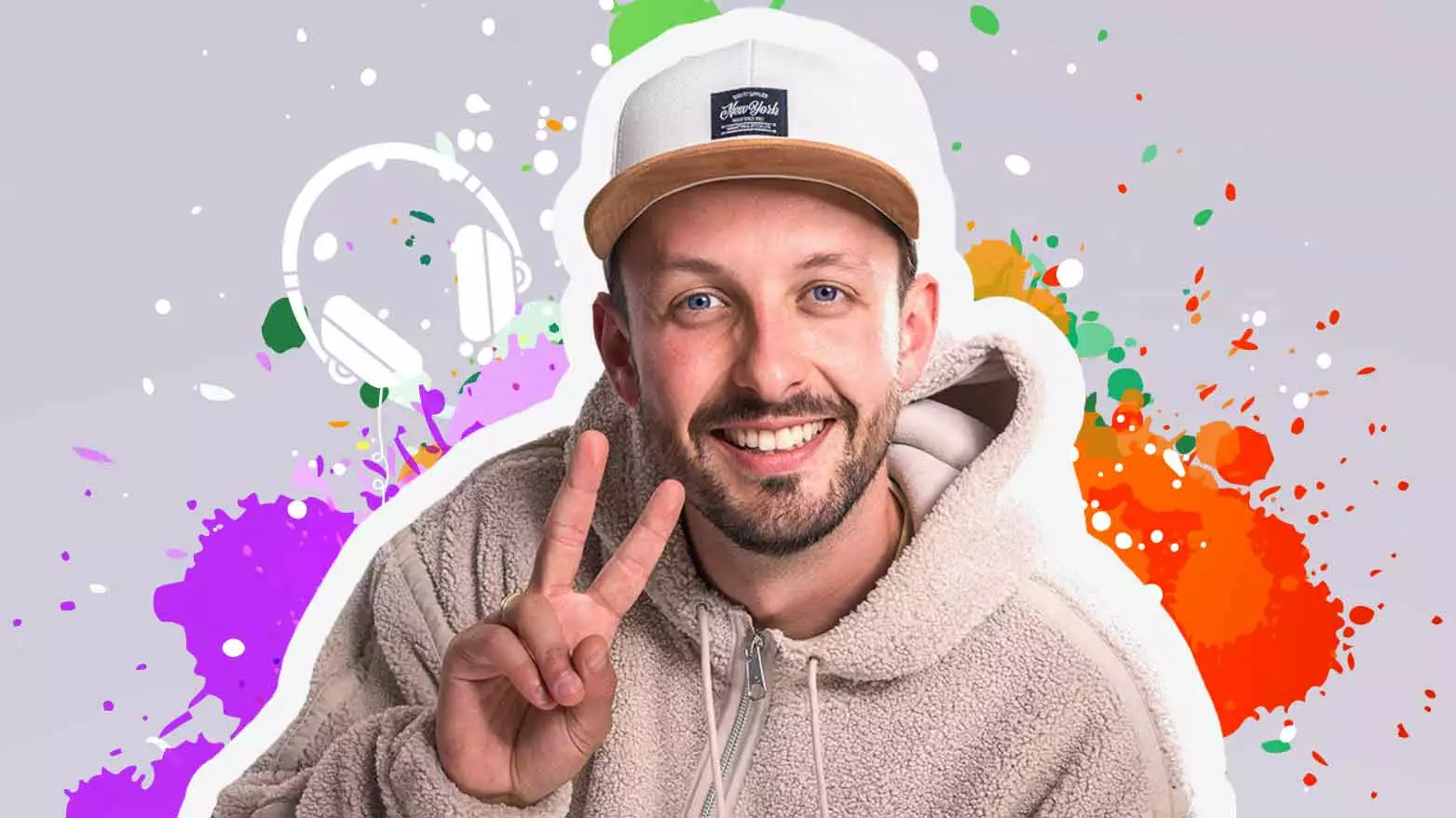 Der Künstler Nielsen blickt freundlich lächelnd in die Kamera. Mit seiner rechten Hand macht er das Peace Zeichen. Er trägt eine beige Baseballcap und eine Kapuzenjacke aus beigem Teddyfell. Auf dem Hintergrund des Bildes sind rote, grüne und lila Farbtupfer und Farbflecken auf einem beigen Hintergrund abgebildet.