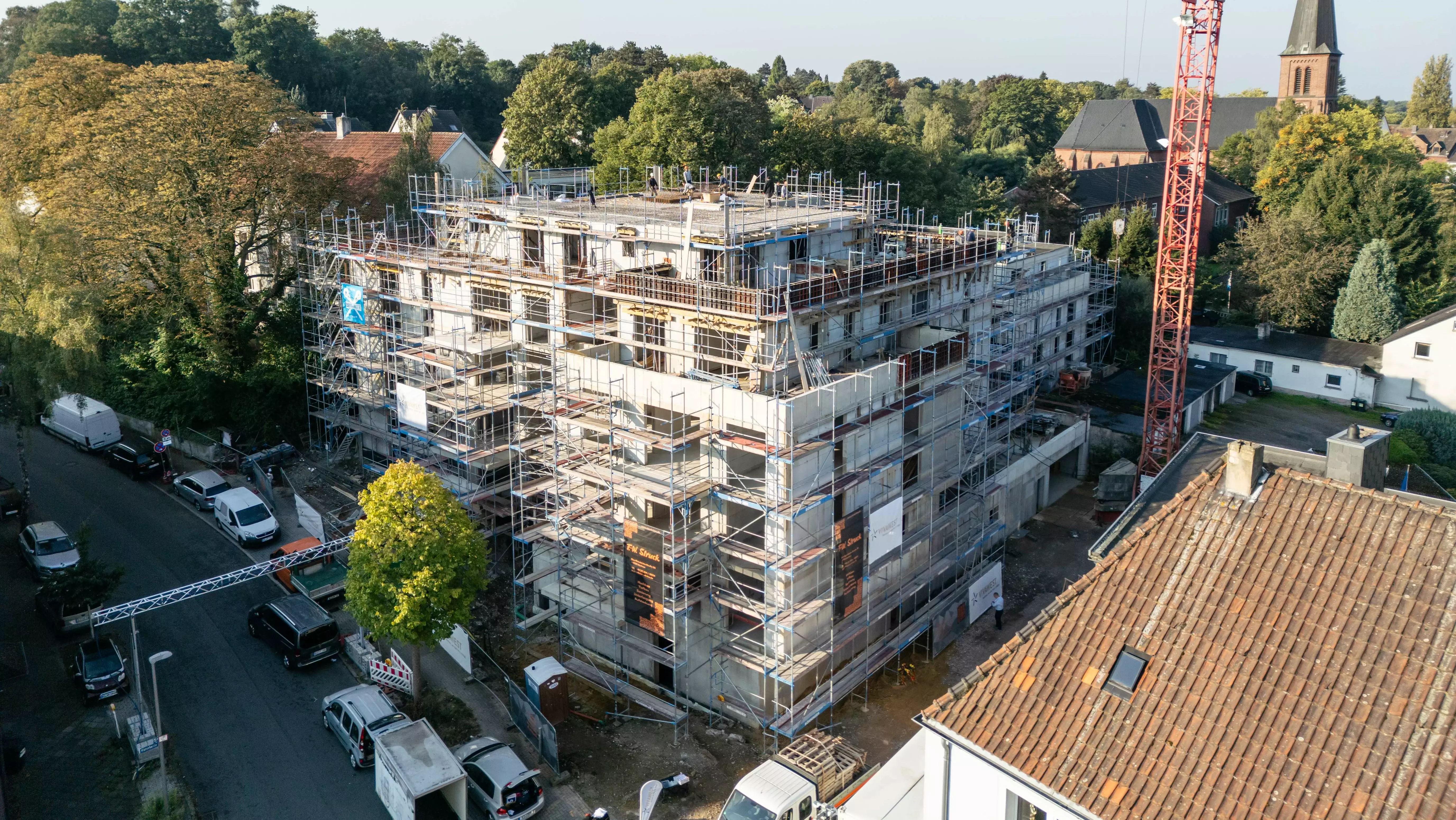 Rohbau, es steht nur der Betonkörper, der von Gerüsten umgeben ist. Im Hintergrund sind Bäume und ein Kirchturm zu sehen.
