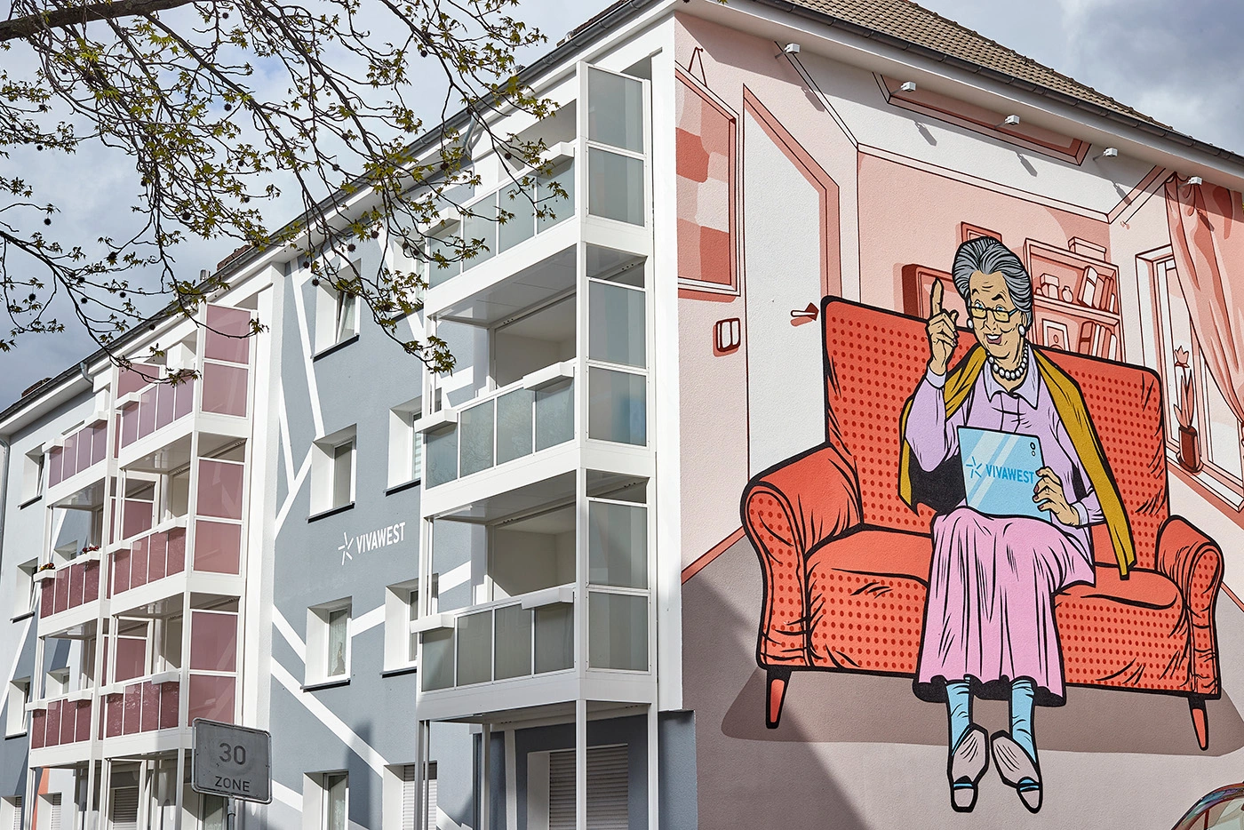 Ein modernisiertes Gebäude. An eine Hauswand wurde ein großes Bild gemalt. Darauf zu sehen ist eine ältere Dame, die in ihrem Wohnzimmer auf einem roten Sofa sitzt und ein Tablet mit VIVAWEST-Schriftzug in der Hand hält