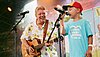 Kinderliedermacher Volker Rosin steht mit einem Kind auf der Bühne des VIVAWEST Family Festival im Kölner Tanzbrunnen. Er hält eine Gitarre in der Hand und blickt lächelnd auf den Jungen neben sich. Der Junge trägt ein Festival-T-Shirt und singt ins Mikrophon.  