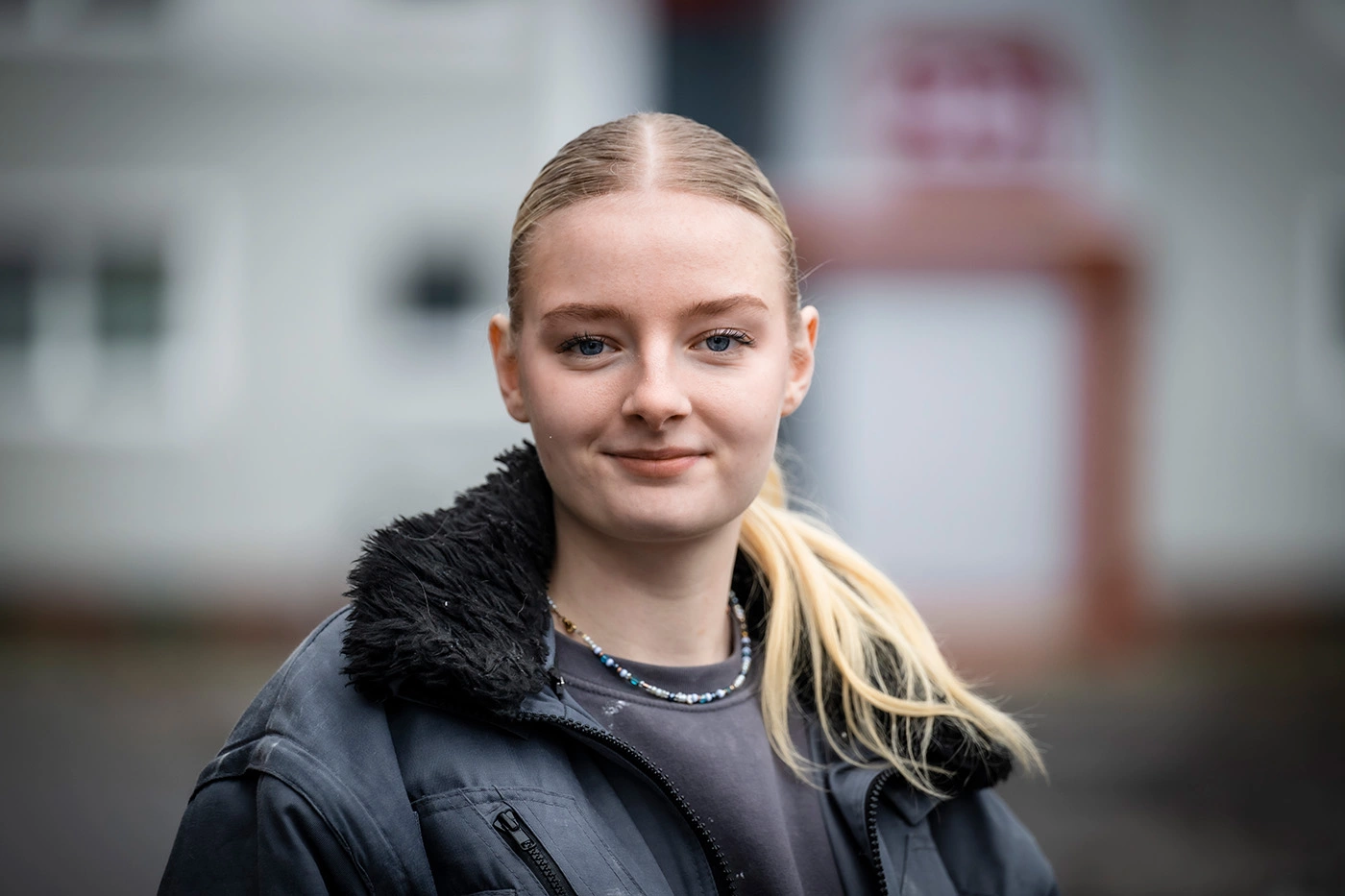 Porträt einer jungen Frau, die direkt in die Kamera schaut und leicht lächelt. Ihre blonden Haare trägt sie in einem Pferdeschwanz. Sie hat einen schwarzen Pulli und eine schwarze Bauarbeiter-Jacke an.