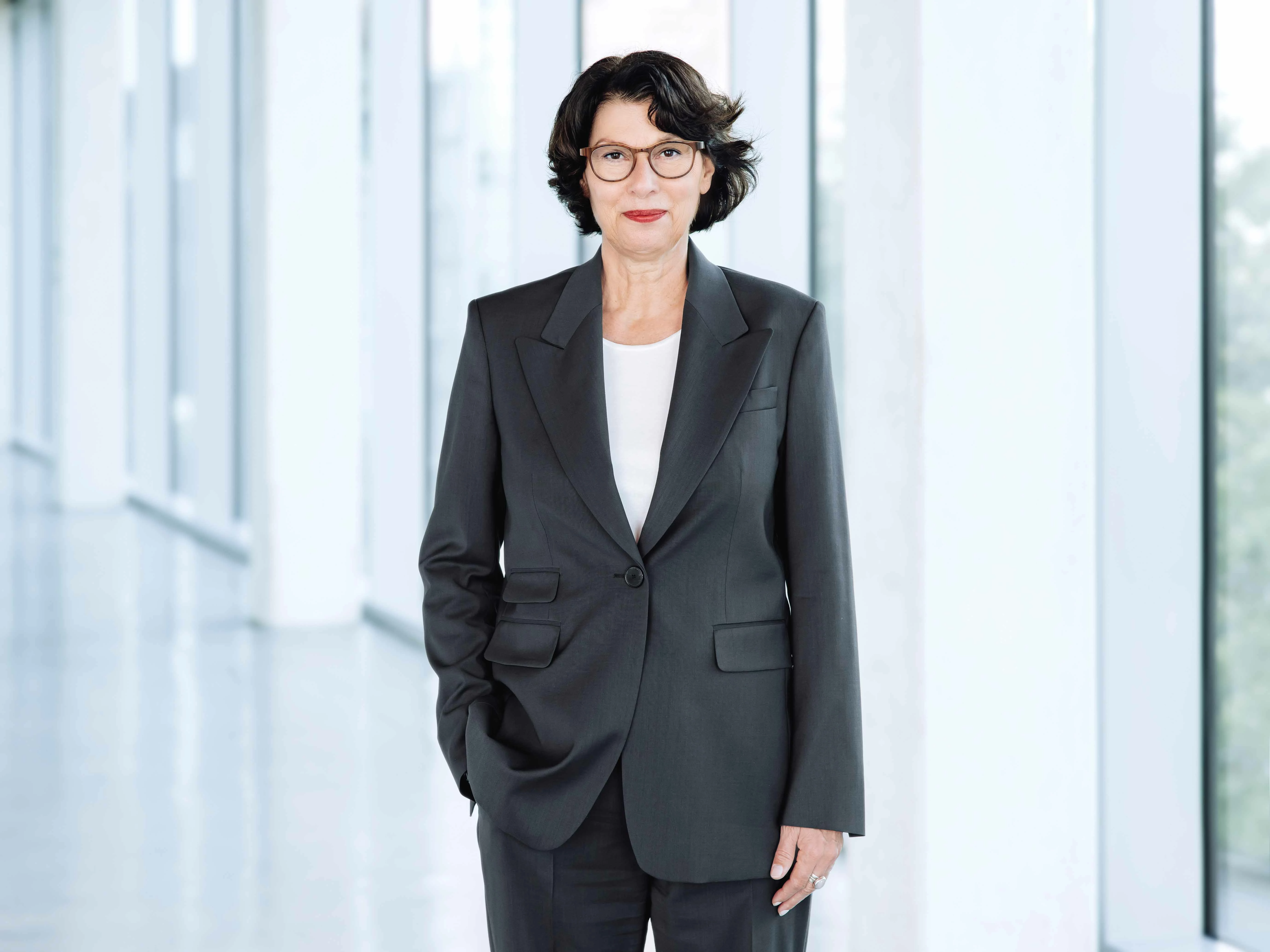 Arbeitsdirektorin Karin Erhard im Portrait. Sie steht vor einer Fensterfront im Foyer des Kesselhauses am VIVAWEST-Campus und schaut lächelnd in die Kamera. Sie trägt einen dunkelgrauen Hosenanzug und eine weiße Bluse.
