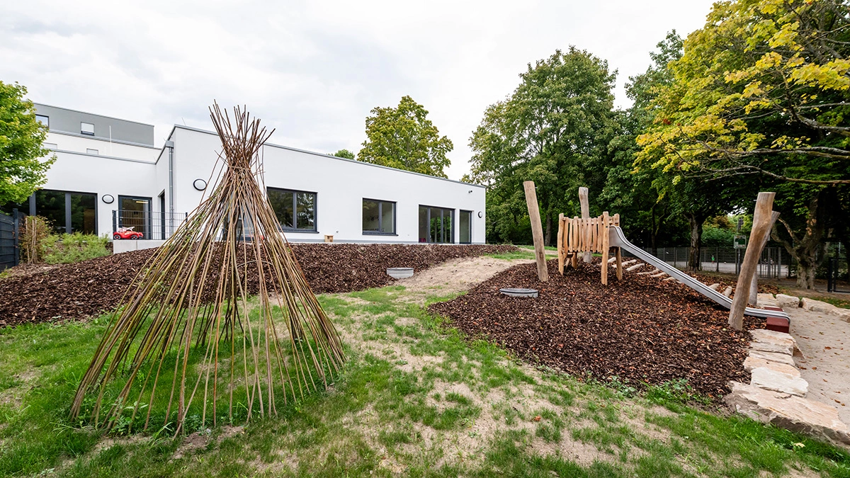 Es ist ein Spielplatz zu sehen mit einem Hügel. Auf diesem befindet sich eine silberne Rutsche und ein aus dünnen Stöcken erstelltes Tipi. Im Hintergrund ist ein weißes Gebäude zu sehen