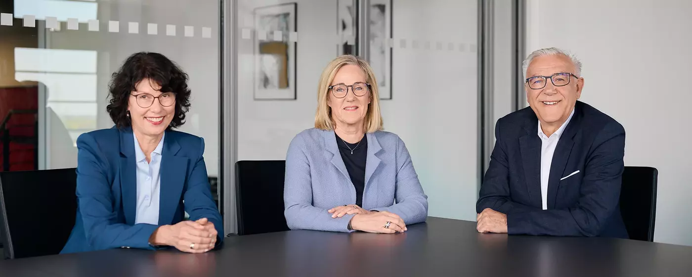Zwei Frauen und ein Mann sitzen an einem schwarzen Bürotisch und lächeln in die Kamera. Hinter ihnen ist eine Glasfront zu sehen.