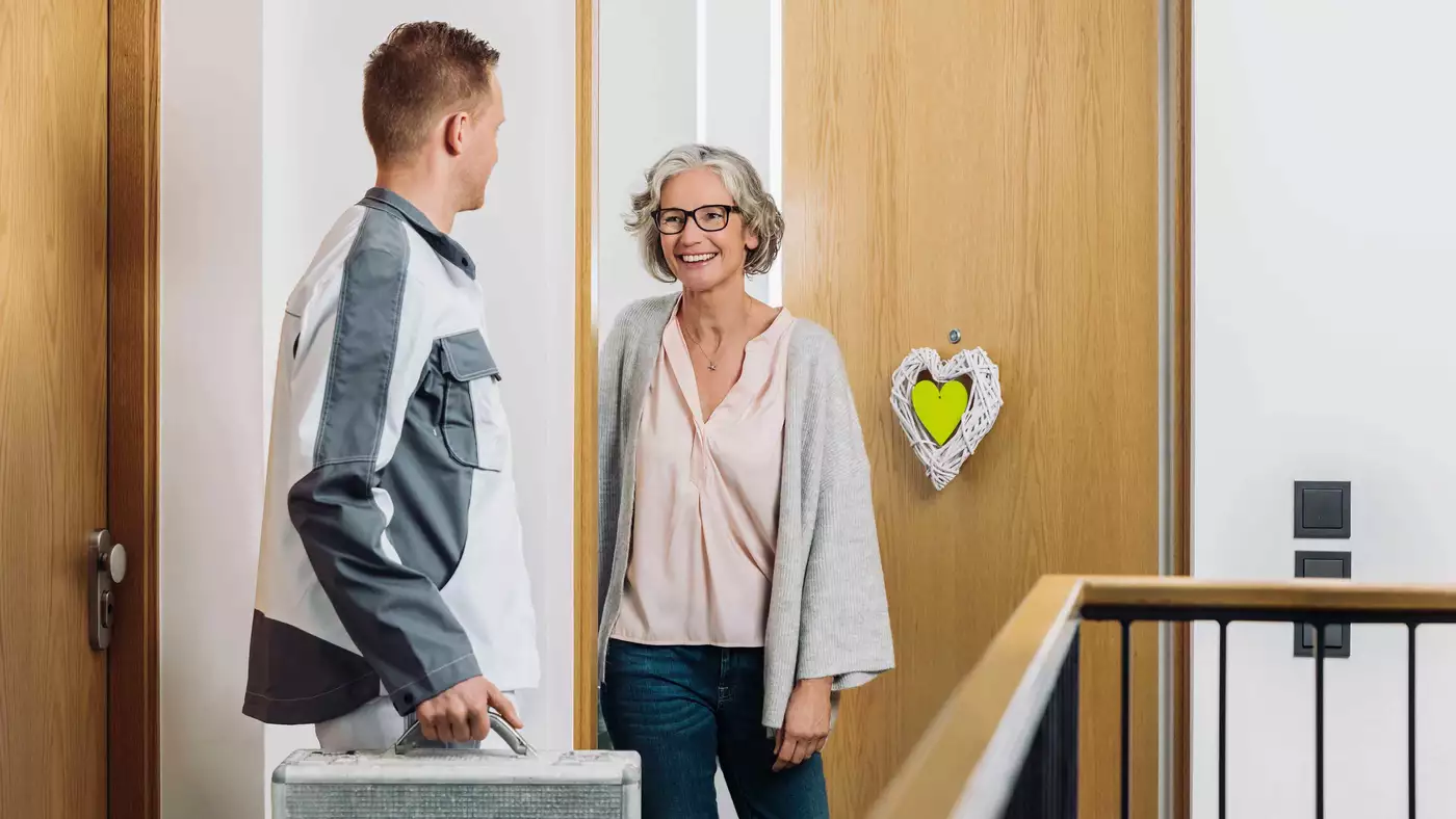 Ein VIVAWEST-Mitarbeiter in weiß-grauer Arbeitskleidung steht mit einem silbernen Koffer in der Hand vor der Wohnungstür einer Mieterin. Die Mieterin hat die Tür geöffnet und blickt ihn lächelnd an.
