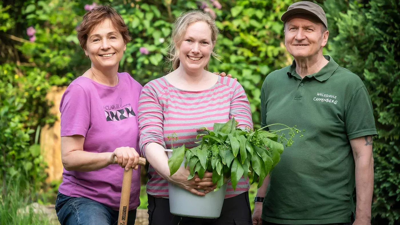 Zwei Frauen und ein Mann stehen in einem Garten. Eine Frau hält einen Griff eines Spatens in der Hand, die zweite Frau hält einen hellblauen Blumentopf mit einer Grünpflanze darin in den Armen. Alle drei schauen in die Kamera und lächeln.