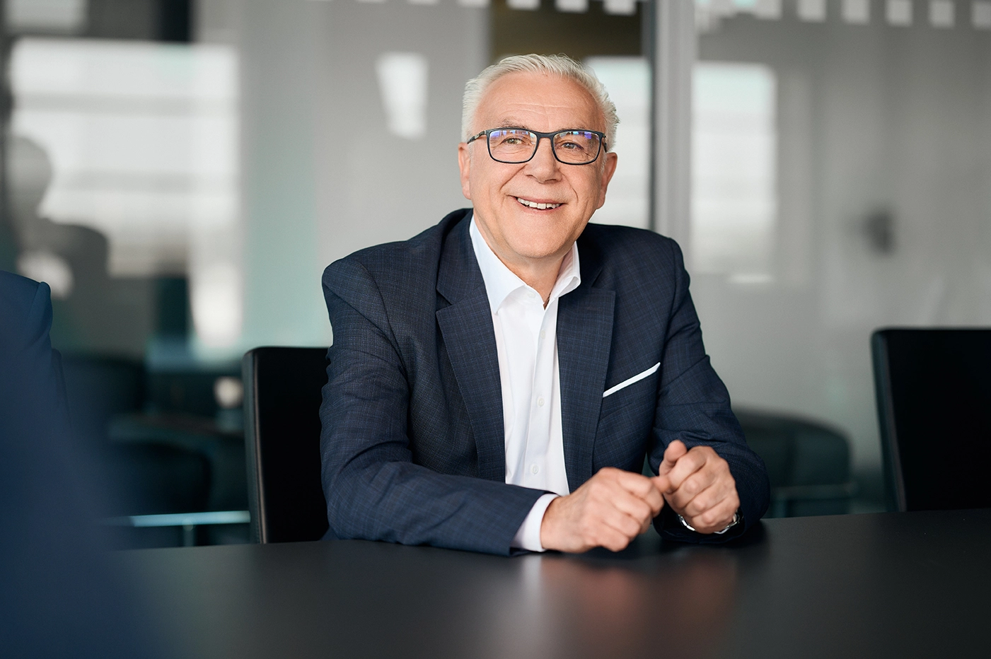 Ein Mann mit grauen Haaren und einer Brille sitzt an einem schwarzen Tisch und lächelt. Sein Blick geht an der Kamera vorbei. Er trägt ein weißes Hemd und ein dunkelblaues Sakko